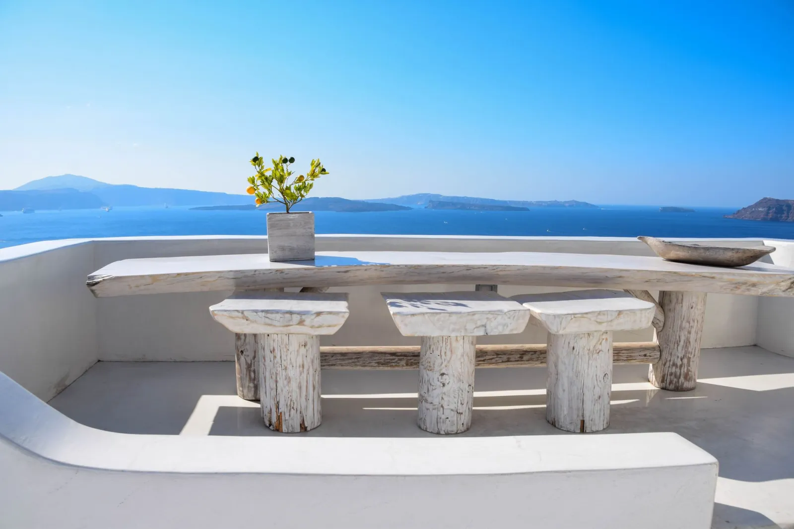 A whitewashed terrace with a wooden table and stools above the Aegean Sea